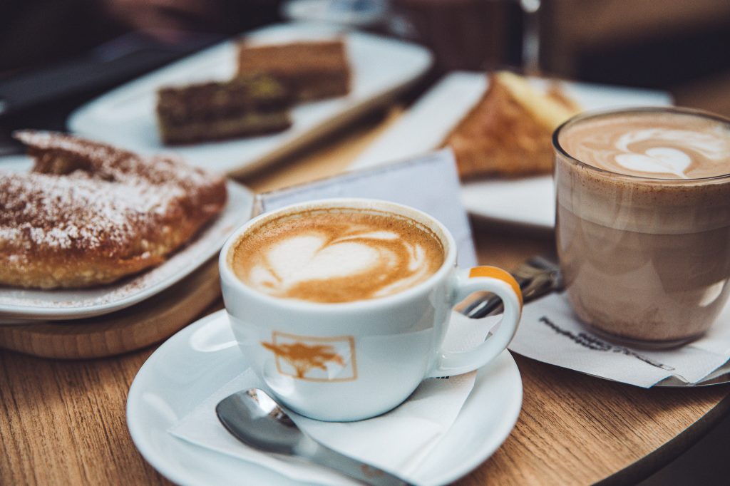 Zwei Tassen Kaffee mit Latte Art auf einem Holztisch, daneben Kuchen und griechische Süßspeisen bei GLYKO in Luxemburg Stadt.