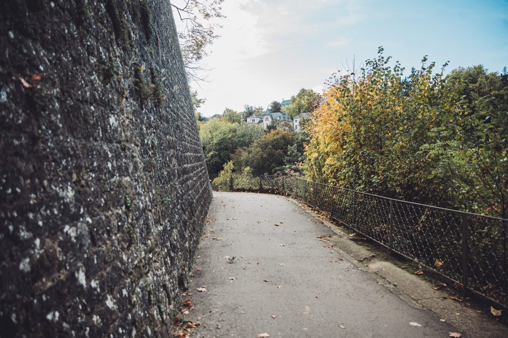 Fußweg in Luxemburg Stadt entlang einer hohen Felswand mit Geländer, Herbstlaub und Bäumen am Rand der Schlucht.