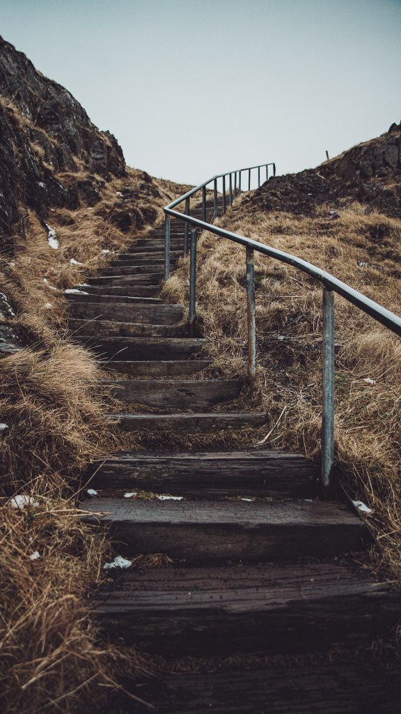 Wooden outdoor staircase with a metal handrail winding up a grassy, rocky hillside.