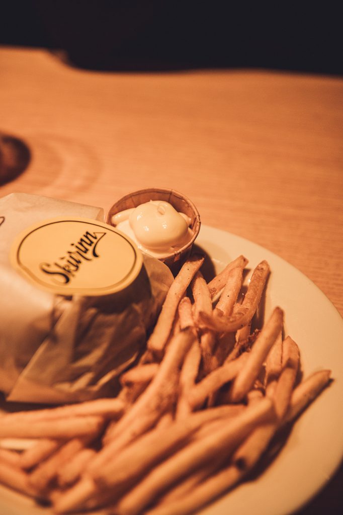 Plate of thin French fries with a small dipping sauce cup and a wrapped item on the side, under warm lighting.
