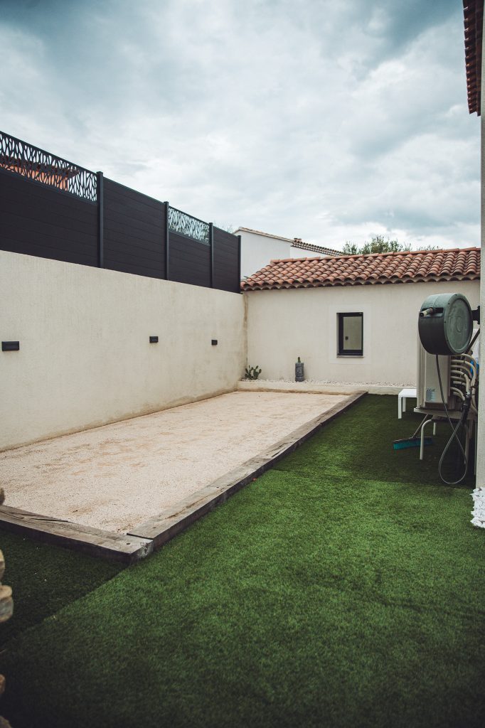 Walled backyard with a beige stucco wall, black decorative fence, and a gravel path next to artificial grass under a cloudy sky