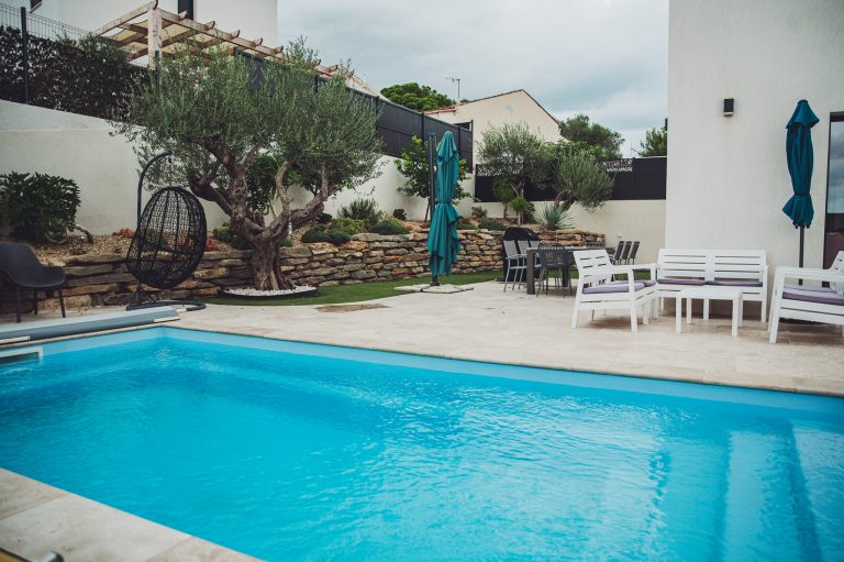 Poolside backyard with bright blue water, stone retaining wall, olive trees, and teal umbrellas over seating area.