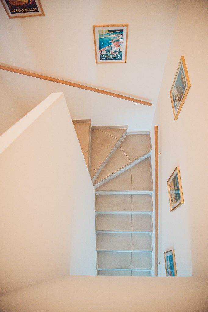 Top-down view of a light stairwell with a wooden handrail and framed posters along the walls.
