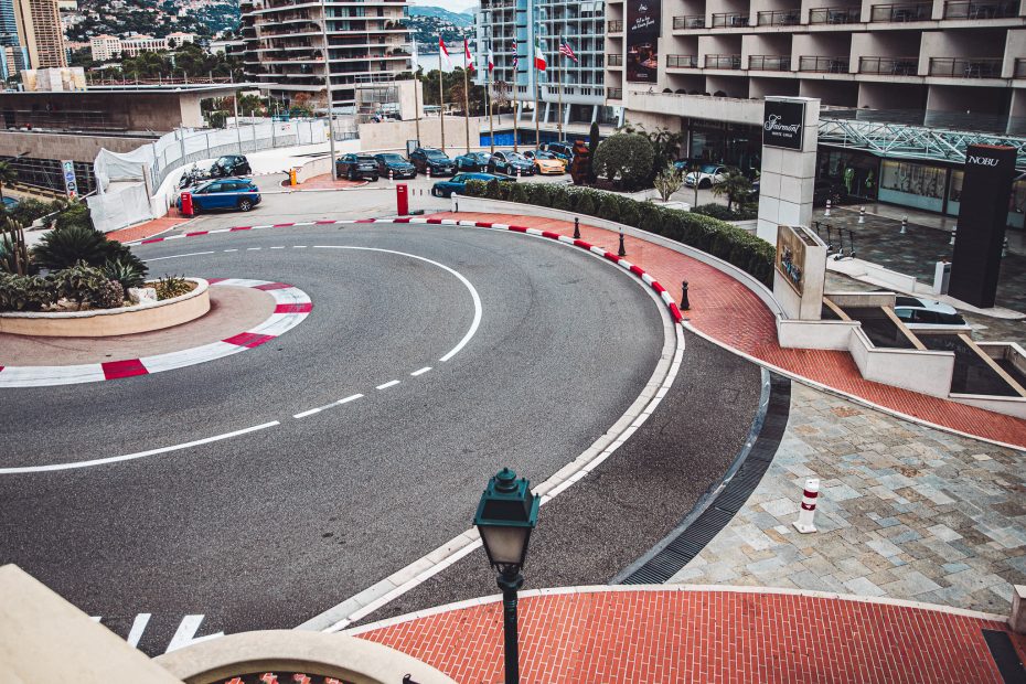 Aerial view of a curved road circling a roundabout in front of a hotel, with flagpoles, parked cars, and red‑and‑white curbs in Monaco.