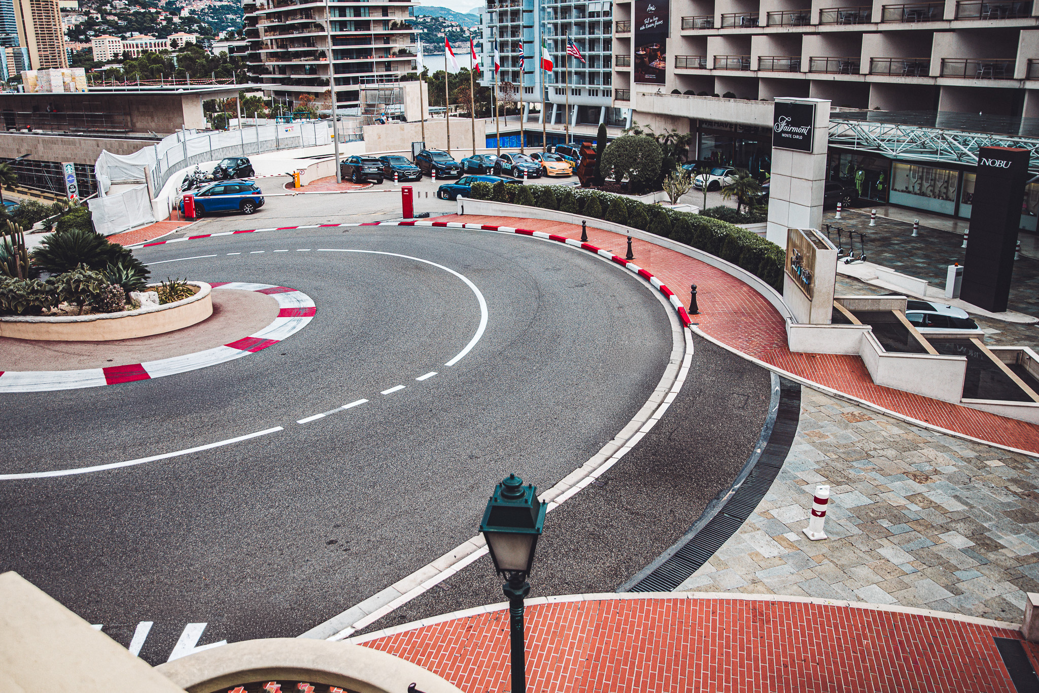 Aerial view of a curved road circling a roundabout in front of a hotel, with flagpoles, parked cars, and red‑and‑white curbs in Monaco.