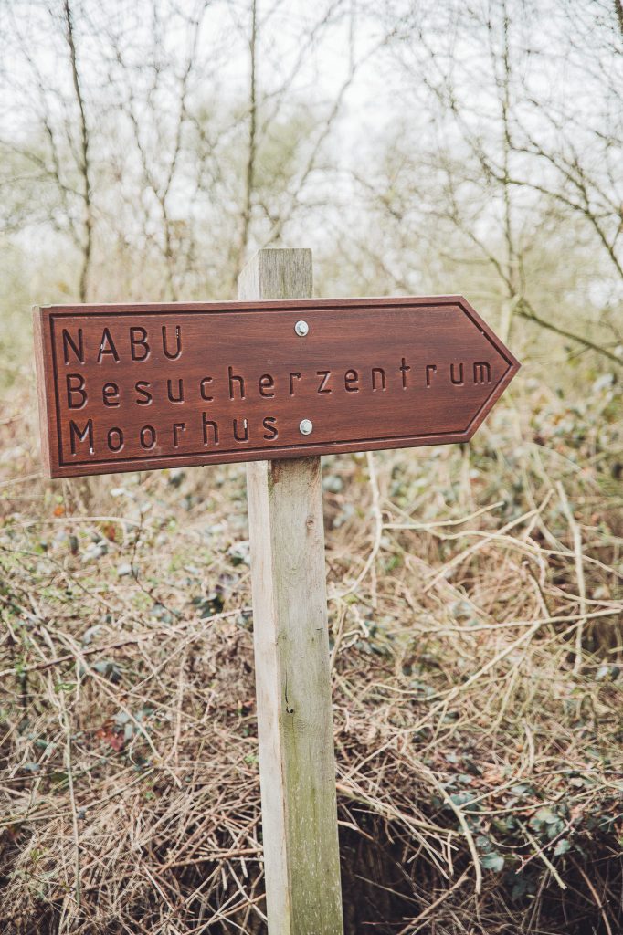 Hölzerner Wegweiser zum NABU Besucherzentrum Moorhus zwischen kahlen Sträuchern