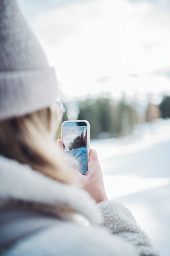Person mit grauer Strickmütze hält ein Smartphone vor eine verschneite Landschaft mit Nadelbäumen