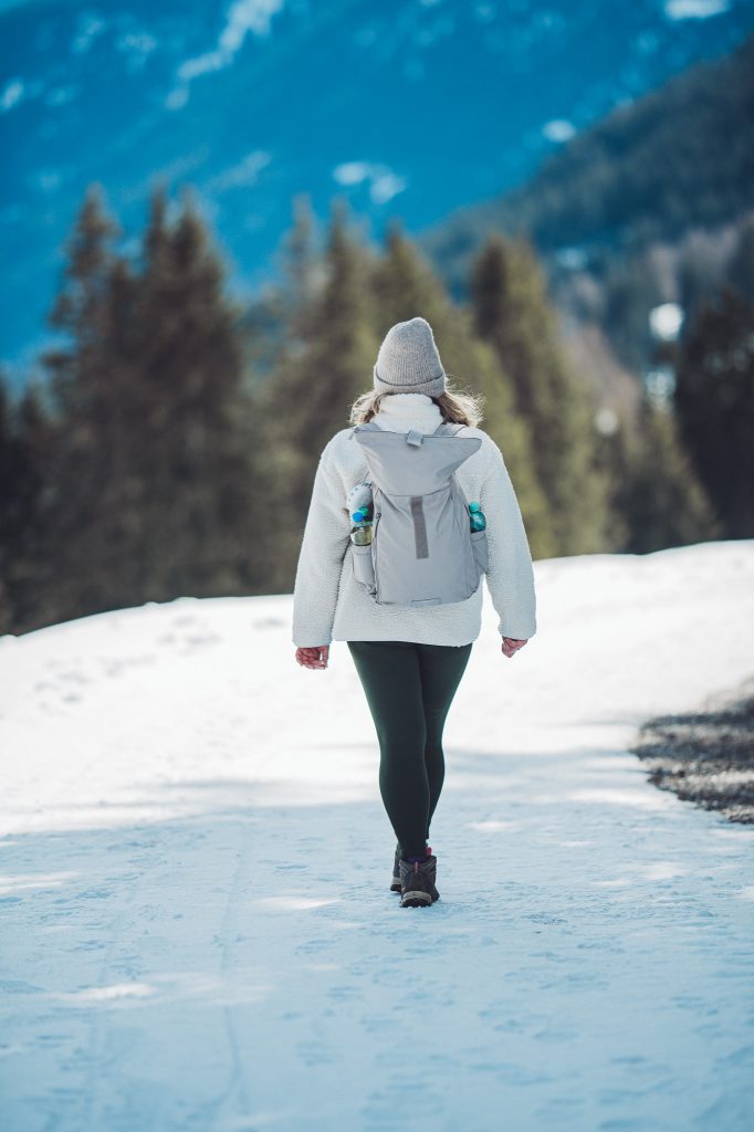 Frau mit grauer Mütze, heller Jacke und Rucksack geht auf einem schneebedeckten Weg in den Alpen