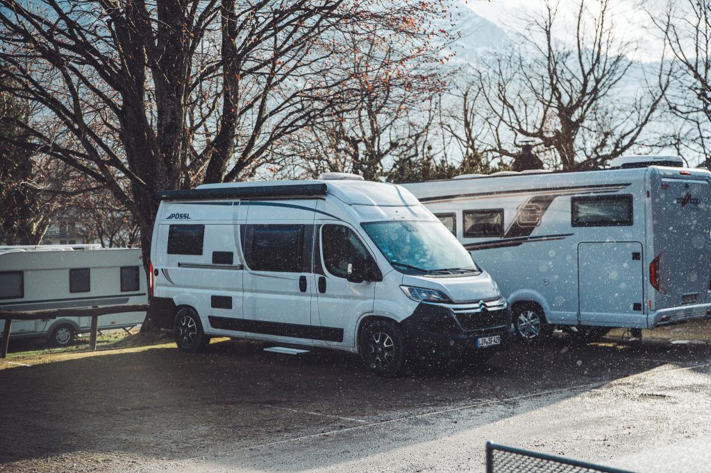 Weißer Kastenwagen neben einem Wohnmobil und einem Wohnwagen unter kahlen Bäumen auf dem Campingplatz Alpenblick bei Interlaken.