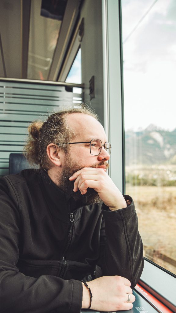 Mann mit Brille und Bart sitzt am Zugfenster und blickt auf die Landschaft draußen