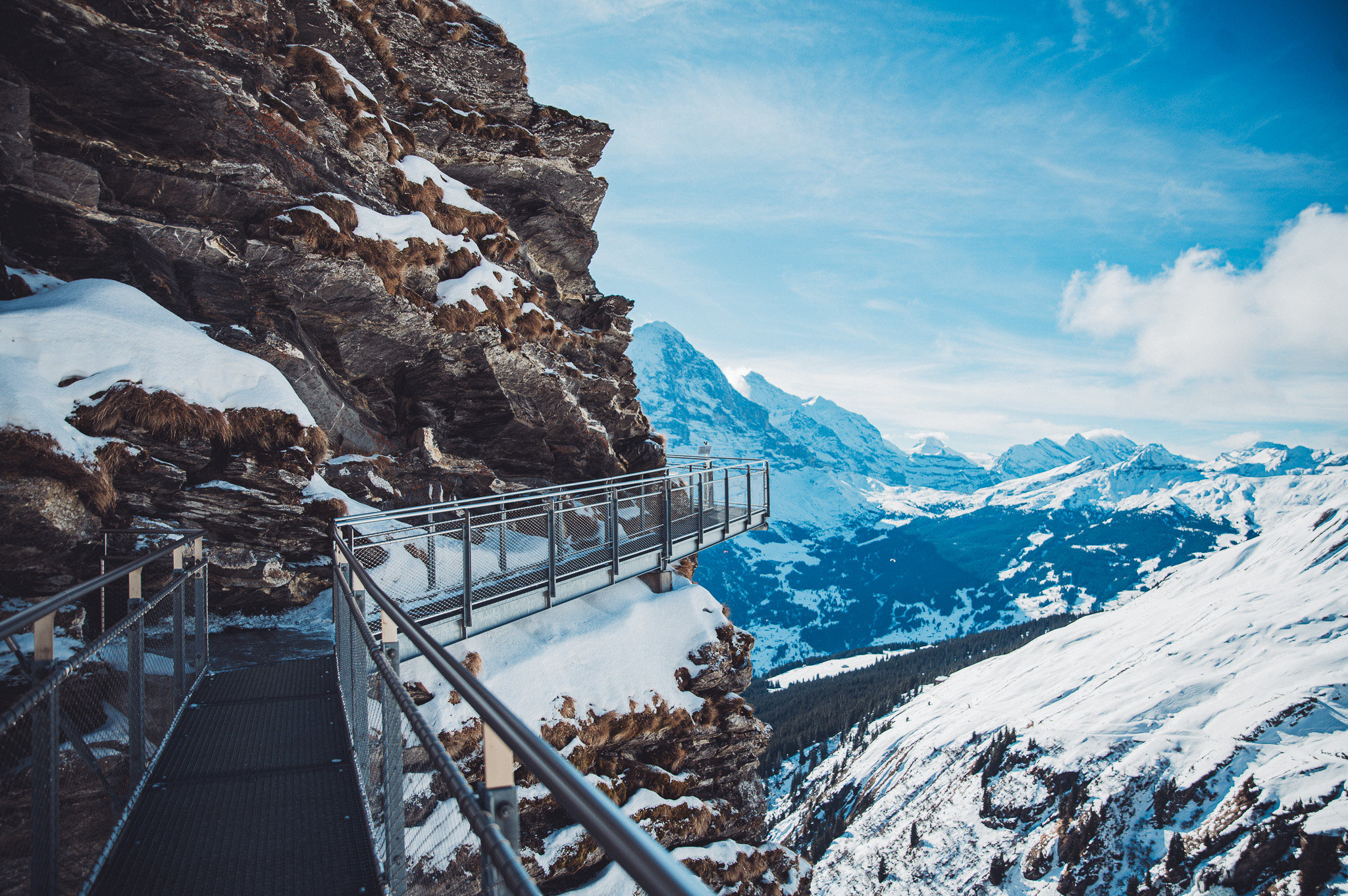 Metallsteg mit Gittergeländer an einer Felswand am First Cliff Walk vor verschneitem Bergpanorama