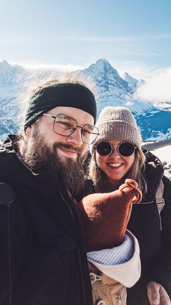 Zwei Erwachsene und ein Baby mit roter Mütze in einer Trage vor verschneiten Bergen am First bei Grindelwald