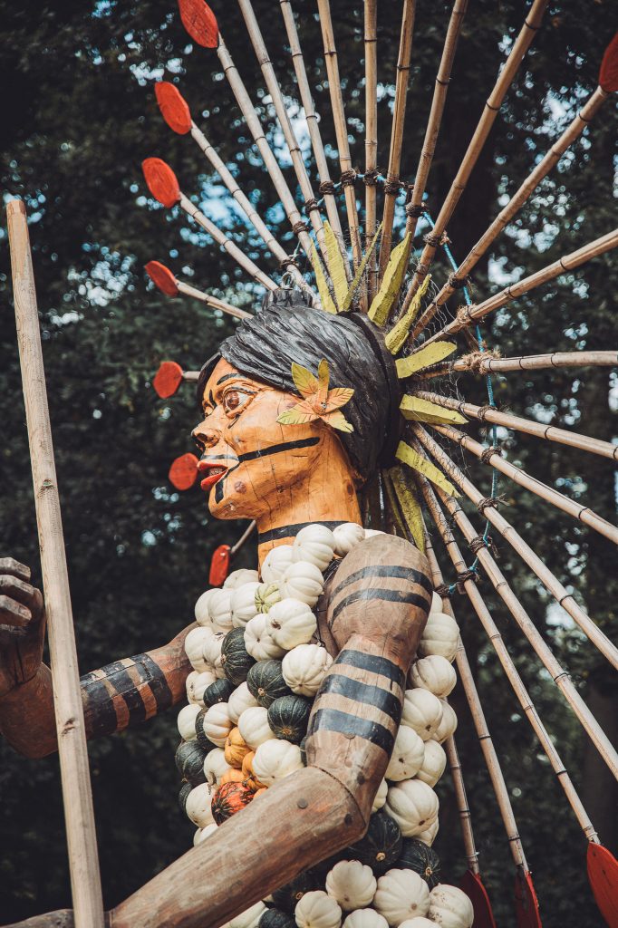 Nahaufnahme einer Holzfigur mit Federschmuck, Stab und einem Kleid aus weißen und grünen Kürbissen